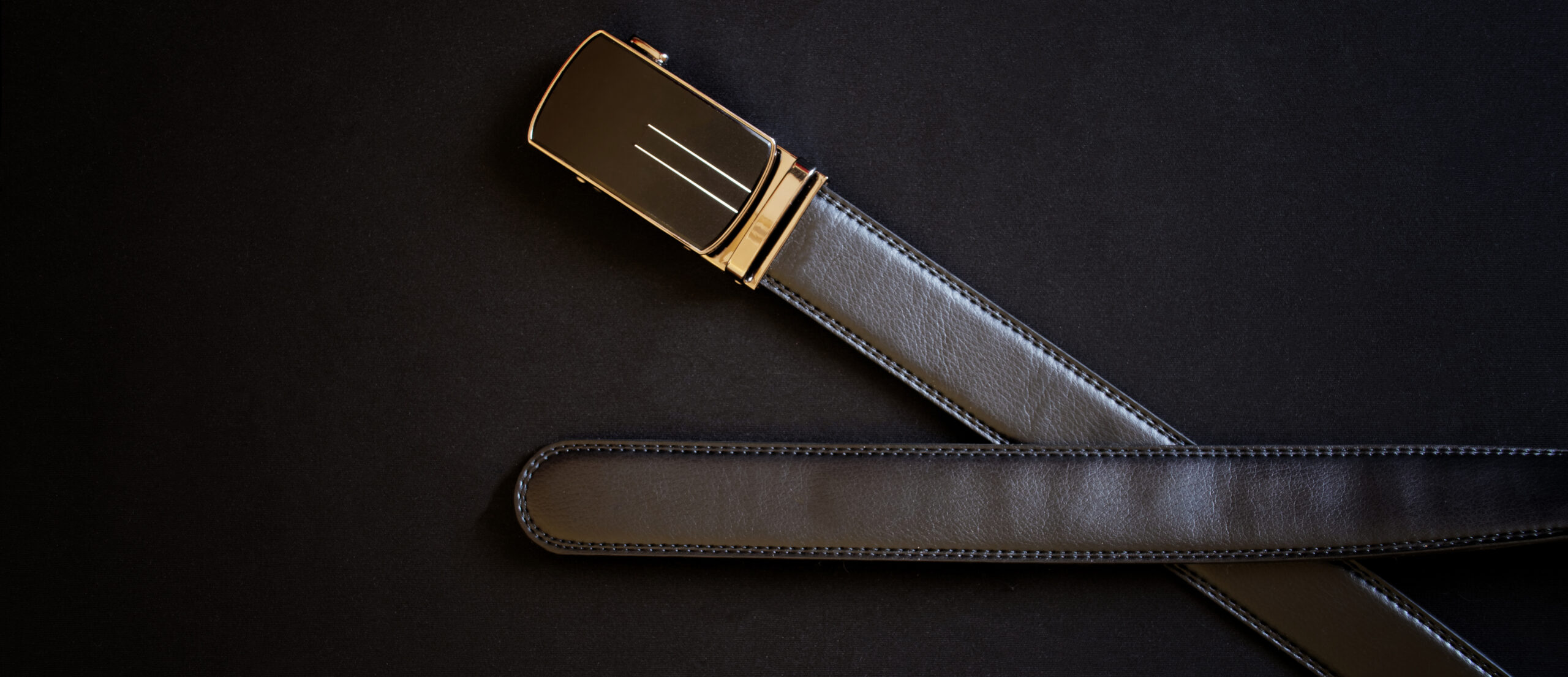 A men's black leather belt with an automatic golden buckle lies against a dark background. Close-up